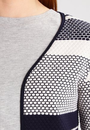Nahaufnahme einer Person, die ein graues Hemd unter einer marineblauen und weißen Strickjacke mit einem Wabenmuster und schwarzem Rand trägt.