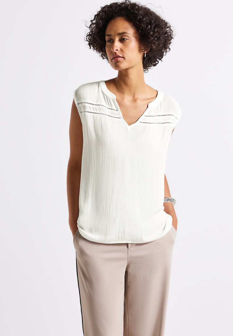 White, textured sleeveless blouse with a V-neck and decorative stitching across the chest, paired with light beige trousers.