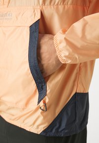 Chaqueta ligera de color durazno con acentos en azul marino. Cuenta con puños elásticos, un bolsillo y un gancho para sujeción. Material liso y resistente al agua.