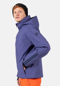 Fioletowa wodoodporna kurtka z kapturem, zapinana na zamek, z czarnymi akcentami. Posiada boczne kieszenie i kontrastowe paski na rękawach.