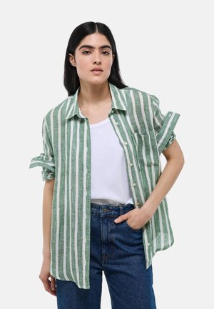 Chemise à rayures vertes et blanches, en tissu léger, manches courtes retroussées, poche avant, portée sur un t-shirt blanc, coupe décontractée.