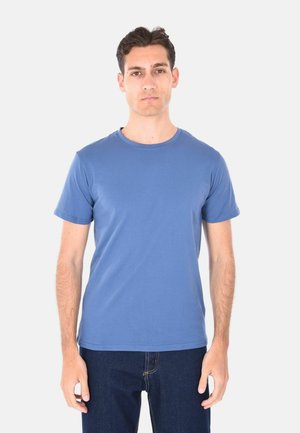 Giovane uomo con capelli castani corti che indossa una semplice maglietta blu a maniche corte e jeans scuri, in piedi davanti a uno sfondo chiaro uniforme.