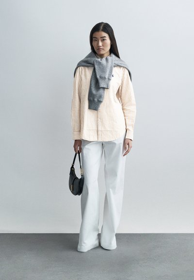 Prúžkovaná košeľa s dlhým rukávom v svetlo oranžovej a bielej farbe, sivá mikina prehodená cez ramená, široké biele nohavice a malá čierna kabelka.