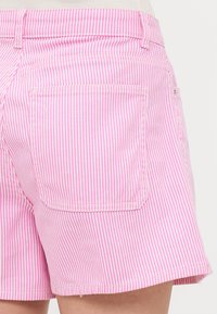 Pantalones cortos a rayas verticales en rosa y blanco con un bolsillo trasero y un dobladillo ligeramente deshilachado, hechos de una tela ligera.