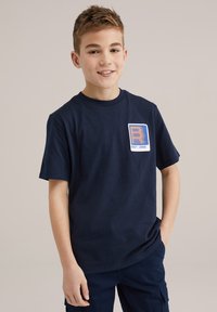 Tamsiai mėlynas medvilninis marškinėliai su oranžine ir mėlyna grafine logo ant krūtinės. Trumpomis rankovėmis ir apvaliu kaklu.