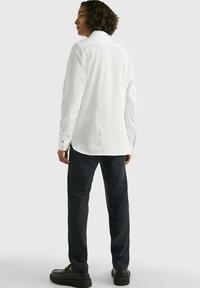 Camisa blanca de manga larga con cuello abotonado, mostrada con pantalones negros texturizados y calzado oscuro. La vista posterior resalta el ajuste entallado de la camisa.