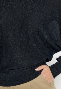 Maglione nero lavorato a maglia con finitura testurizzata e scintillante, vestibilità ampia e polsini e orlo a coste. Mano appoggiata nella tasca di pantaloni color sabbia.