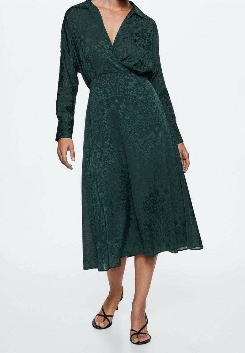 Femme portant une robe portefeuille à manches longues vert foncé avec des motifs floraux subtils et des sandales noires à brides sur fond blanc.