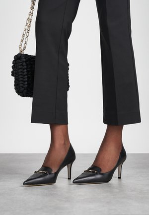 Sapatos de salto alto em pele preta com bico fino e detalhes decorativos em dourado. Combinados com calças pretas estruturadas e uma bolsa texturizada.