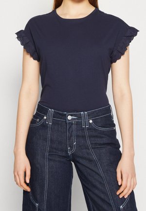 Granatowa koszulka z falbanowymi koronkowymi rękawami, zestawiona z wysokim stanem ciemnych dżinsów z kontrastującymi białymi przeszyciami.