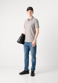 Siva polo majica s kratkimi rokavi, modri jeans, črne superge in črna kapa z napisalom "BOSS" v dvignjenem tisku, drži črno torbo.