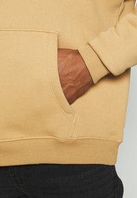 Felpa beige con cappuccio, tasca a marsupio, polsini e orlo a costine. Il tessuto appare morbido con una texture liscia e dettagli di cuciture sottili.