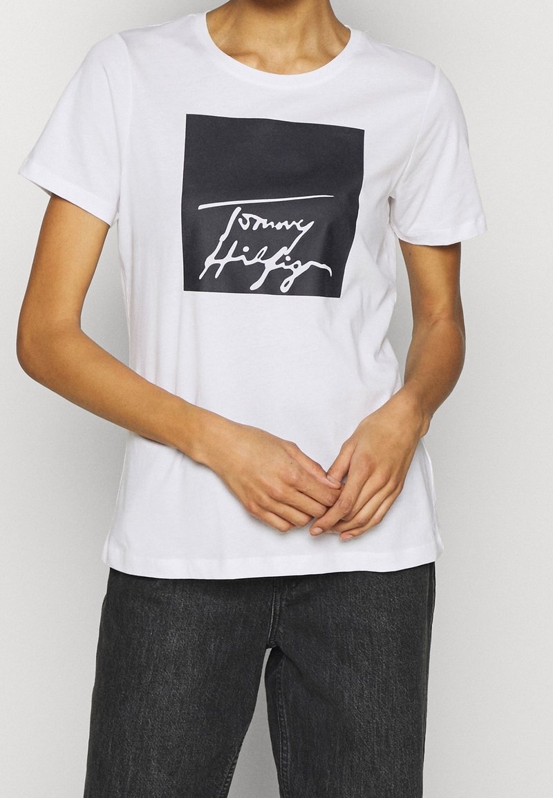 Bílý bavlněný tričko s krátkými rukávy s černým čtvercovým grafickým potiskem a bílým kurzívovým textem "Tommy Hilfiger." Jednoduchý, uvolněný střih.