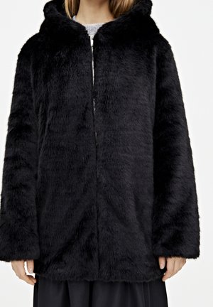 Manteau à capuche en fausse fourrure noire avec fermeture éclair devant, porté sur un pull gris et une jupe noire, visible du cou jusqu'à mi-cuisse.