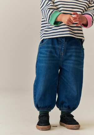 Barn iført stribet langærmet skjorte med grønne og pink manchetter, løse blå jeans med elastiske ankler, sorte sko, stående på en ensfarvet baggrund.