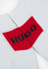 Valkoiset puuvillasukat, joissa on suuri punainen läiskä, jossa on mustalla tekstillä selvästi näkyvissä "HUGO". Karkea neuloskangas, nilkkapituinen malli.