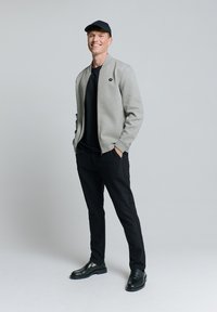 Grijze bomberjack met een zwarte rits, zwart T-shirt, zwarte broek en zwarte schoenen met een gladde textuur. Draag het met een zwarte pet.