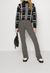 Czarny i szary sweter w wzory z wysokim kołnierzem, zestawiony z szarymi spodniami flare o prążkowanej fakturze. Modelka trzyma małą czarną torbę i nosi buty na grubej białej podeszwie.