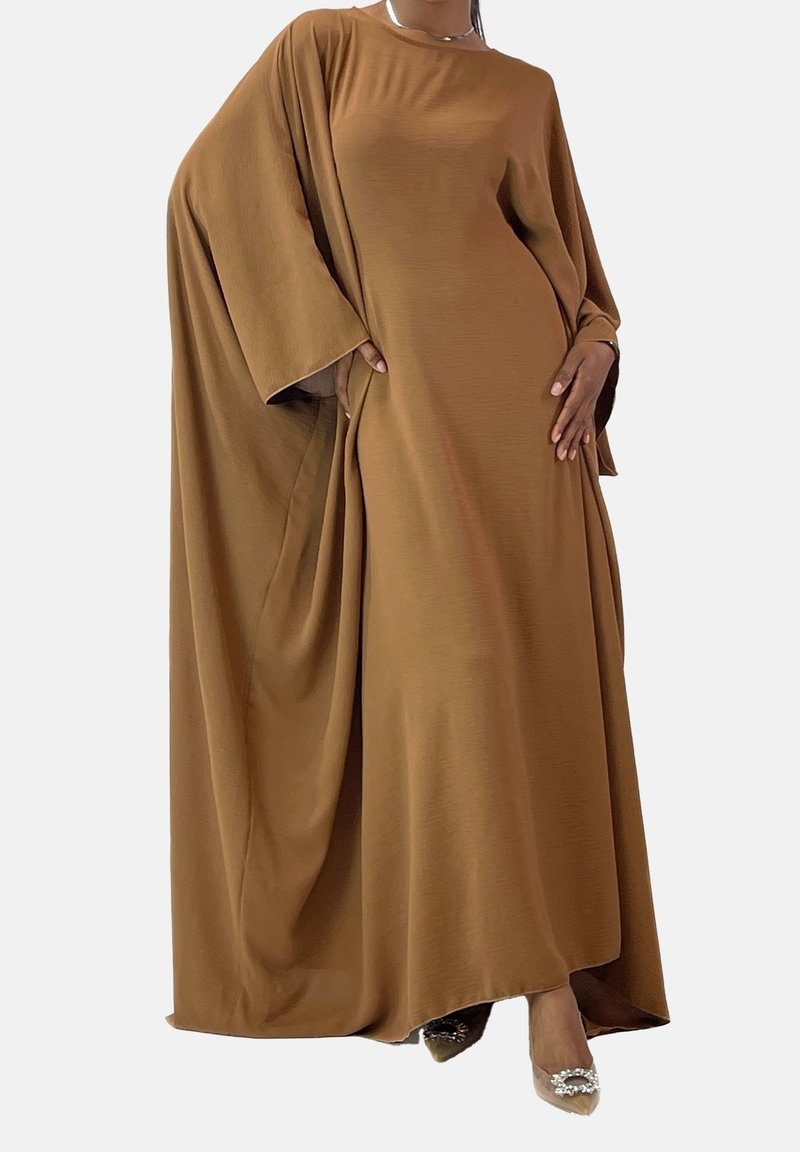 Femme portant une robe longue marron ample avec des manches larges et des talons pointus avec des boucles décoratives sur un fond uni.