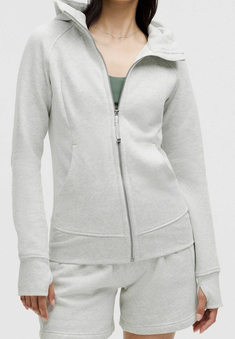 Femme portant un sweat à capuche gris clair zippé avec des trous pour les pouces et un short assorti, montrant le haut du corps et le torse.