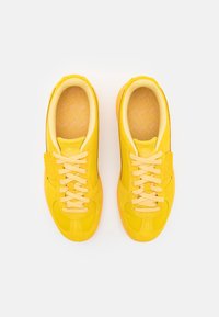Baskets jaunes avec un dessus en cuir lisse, présentant une semelle texturée, un système de laçage et un rembourrage autour du col. Semelle intérieure texturée visible.