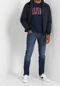 Chaqueta navy, camiseta gráfica azul con texto rojo, jeans de mezclilla oscuros, zapatillas blancas con detalles negros. Atuendo casual con ropa en capas.