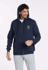 Sweat à capuche zippé bleu marine fabriqué en tissu doux, avec une poche avant et un patch logo sur la poitrine, doté d'une capuche ajustable avec cordon.