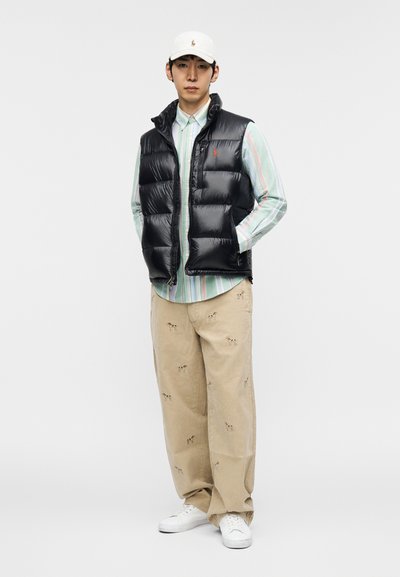 Fekete steppelt mellény többszínű csíkos gombos ing felett, bézs nadrág kutyahímzéssel és fehér sportcipő. A sapka fehér logóval.