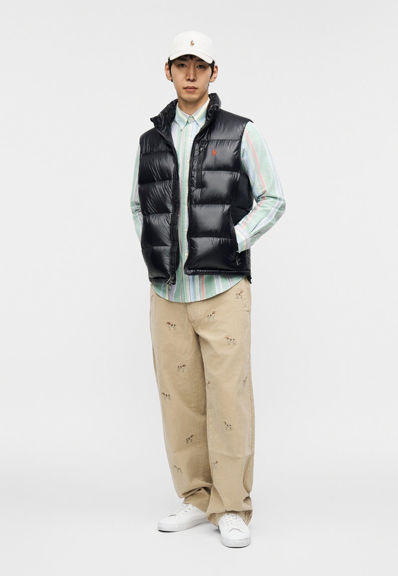 Veste matelassée noire sans manches portée sur une chemise à boutons rayée multicolore, pantalon beige avec broderie de chien, et baskets blanches. Casquette blanche avec un logo.