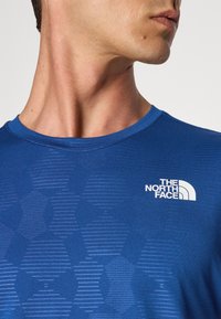 Kék nedvszívó sportpóló rövid ujjakkal, textúrált mintával és fehér logóval a bal mellen. Simasagú szövetfelület.