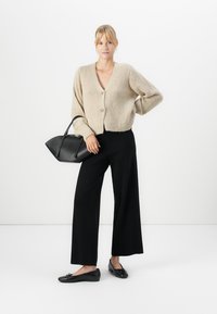 Kvinne iført beige knappevest, svarte vide bukser, svarte leiligheter, holder en svart håndveske med den ene hånden på hoften mot hvit bakgrunn.