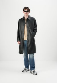 Schwarzer lederner Mantel mit geknöpfter Vorderseite, großem Kragen und Seitentaschen, getragen über einem beigen Hemd und blauen Jeans, kombiniert mit bunten Sneakers.