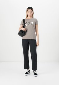 Young woman wearing gray GAP t-shirt, black pants, black sneakers, and holding black shoulder bag against white background.