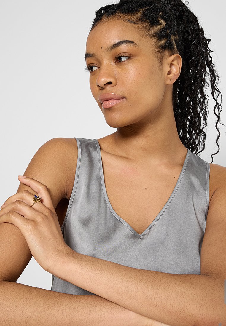 Grau satin Oberteil mit V-Ausschnitt, das eine glatte Textur und sanften Glanz aufweist. Das Model posiert mit verschränkten Armen und trägt einen schlichten Ring an einer Hand.