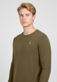 Olivově zelené žebrované tričko s dlouhým rukávem, kulatým výstřihem a jemným logem na hrudi, vyrobené z měkkého, texturovaného materiálu.