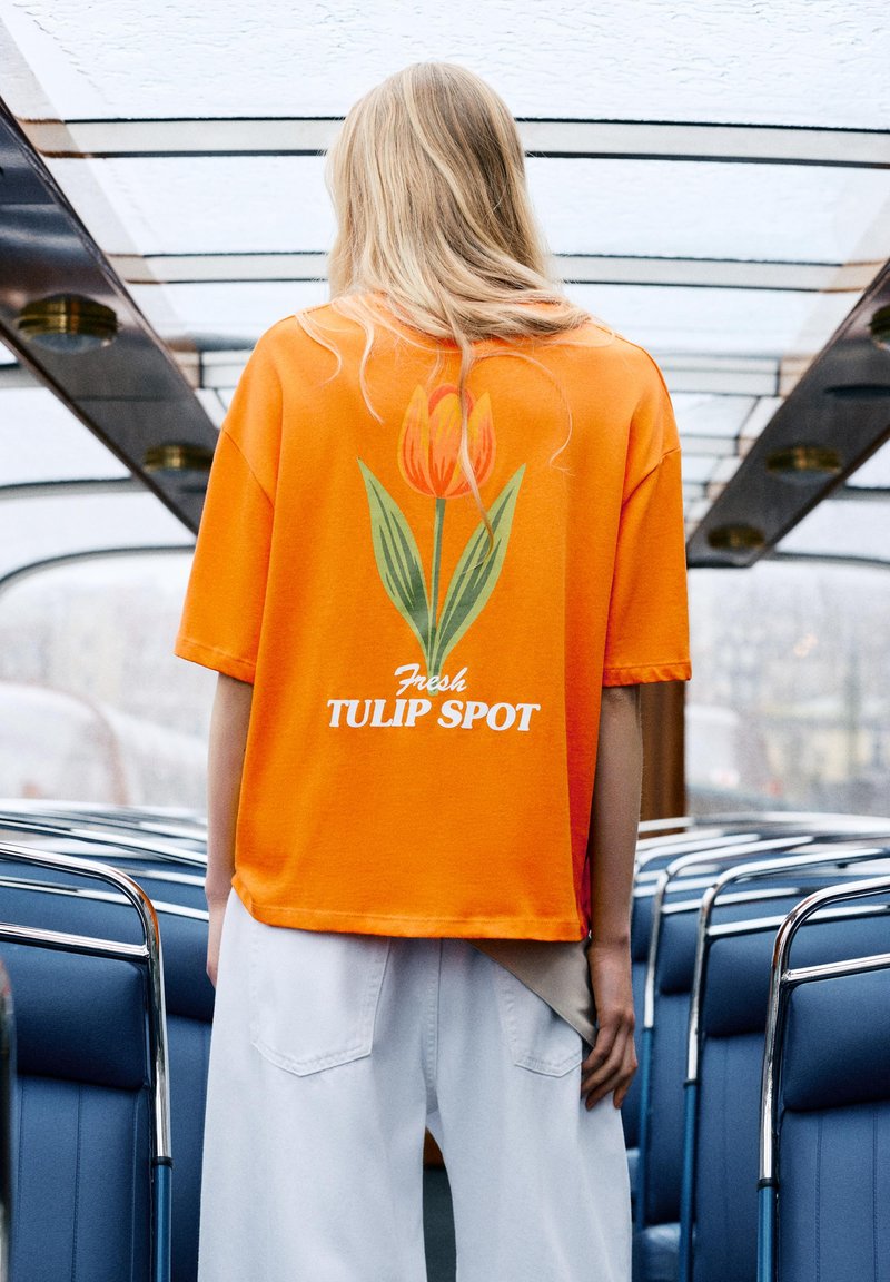 Personne blonde portant un t-shirt orange avec un motif de tulipe et le texte « Fresh Tulip Spot », debout dans l’intérieur d’un bus ou tram à toit en verre.