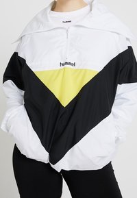 Biała, czarna i żółta kurtka wiatrówka z krótkim zamkiem, dużym kołnierzem, geometrycznym wzorem w blokach kolorów oraz logo Hummel na piersi.