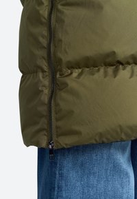 Olijfgroene pufferjack met een zijrits, contrasterende zwarte hardware en een gladde textuur, gecombineerd met blauwe denim jeans.