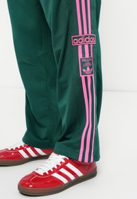 Grønne træningsbukser med pink side-striber og knapper; har et Adidas-logo patch. Parret med røde sneakers og hvide striber.