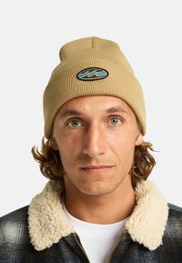 Beanie beige lavorato a maglia con una texture a coste e un logo caratterizzato da motivi ondulati in turchese su una patch nera ovale.