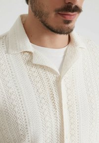 Homme avec une barbe courte portant une chemise en dentelle texturée couleur crème par-dessus un t-shirt blanc à col rond sur un fond neutre.