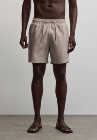 Homme portant un short beige à cordon et des tongs noires, debout sur une surface grise devant un fond gris clair uni.