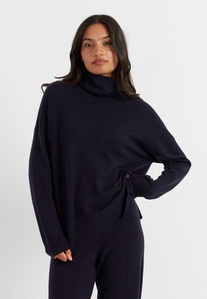 Pull à col roulé bleu marine en tissu doux, présentant une coupe décontractée, des épaules tombantes et des poignets côtelés avec un détail de couture en diagonale.
