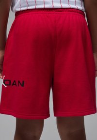 Shorts de sport rouges en matériau léger, avec une ceinture élastique et un logo noir "JORDAN" accompagné d'une silhouette blanche sur la jambe gauche.