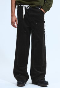 Person, der lockere schwarze Cordhosen mit großen vorderen Taschen, schwarze Schuhe und ein grünes Langarmshirt trägt, mit einer Hand in der Tasche.