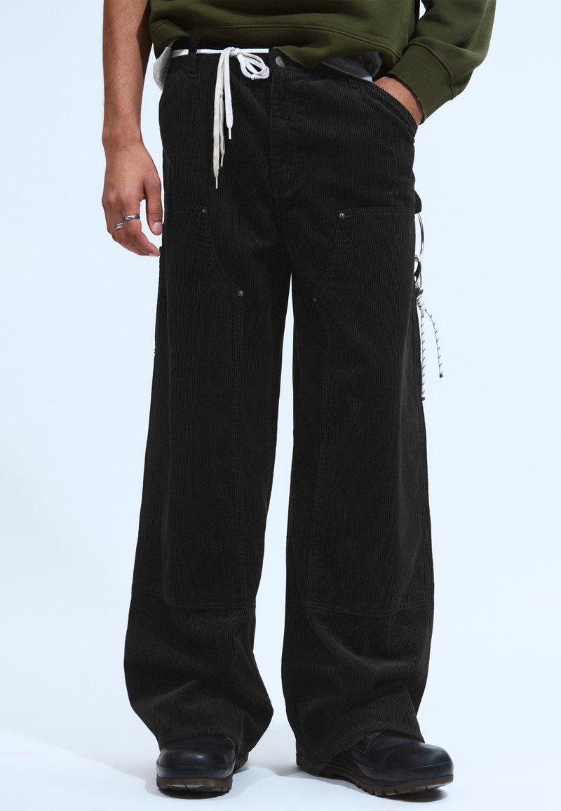 Person, der lockere schwarze Cordhosen mit großen vorderen Taschen, schwarze Schuhe und ein grünes Langarmshirt trägt, mit einer Hand in der Tasche.