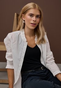 Blondinė moteris su perlų auskarais, vilkinti baltą atvirą palaidinę ant juodo viršaus ir tamsių kelnių, sėdinti prieš rudos spalvos foną.