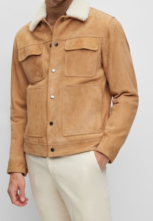 Tan suede jacket with white shearling collar, front flap pockets, and snap buttons worn over a brown turtleneck and cream pants.