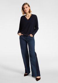 Marineblauer V-Ausschnitt-Pullover mit langen Ärmeln, kombiniert mit dunkelblauen, weit geschnittenen Jeans mit kontrastierenden Seitenteilen; braune spitze Ballerinas vervollständigen das Outfit.