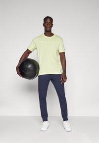 Lichtgroen T-shirt met korte mouwen met het "Champion" logo, gecombineerd met marineblauwe broek en witte sneakers, terwijl hij een zwarte medicijnbal vasthoudt.